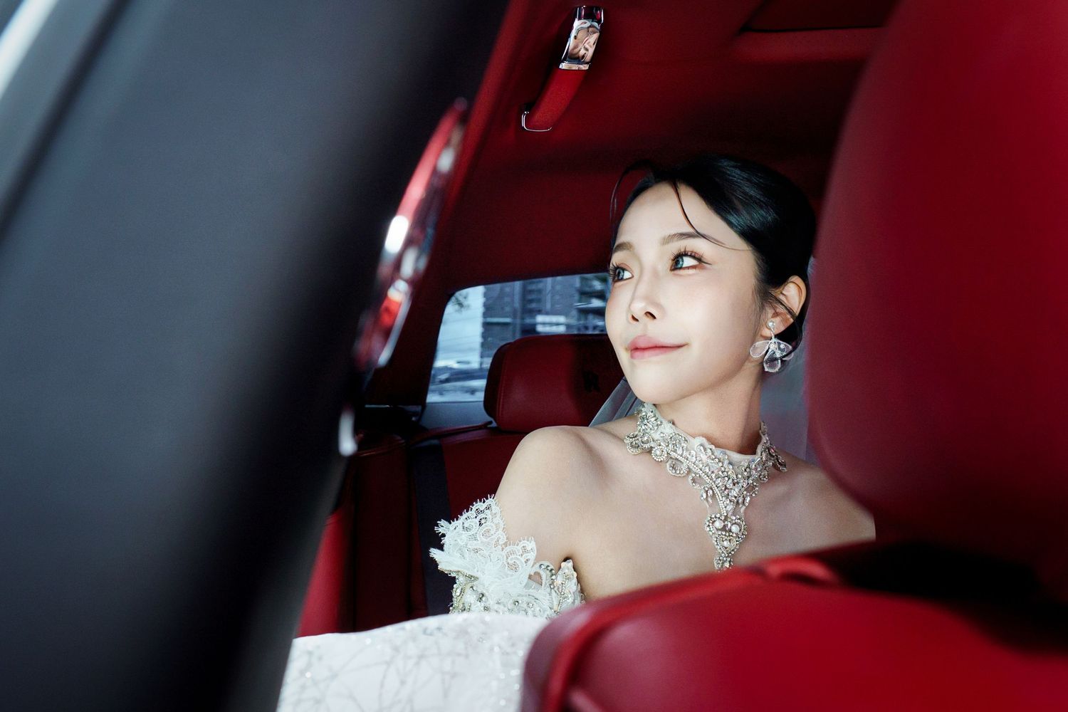 A bride wearing a white wedding dress and sparkling earrings sits in a red car interior.