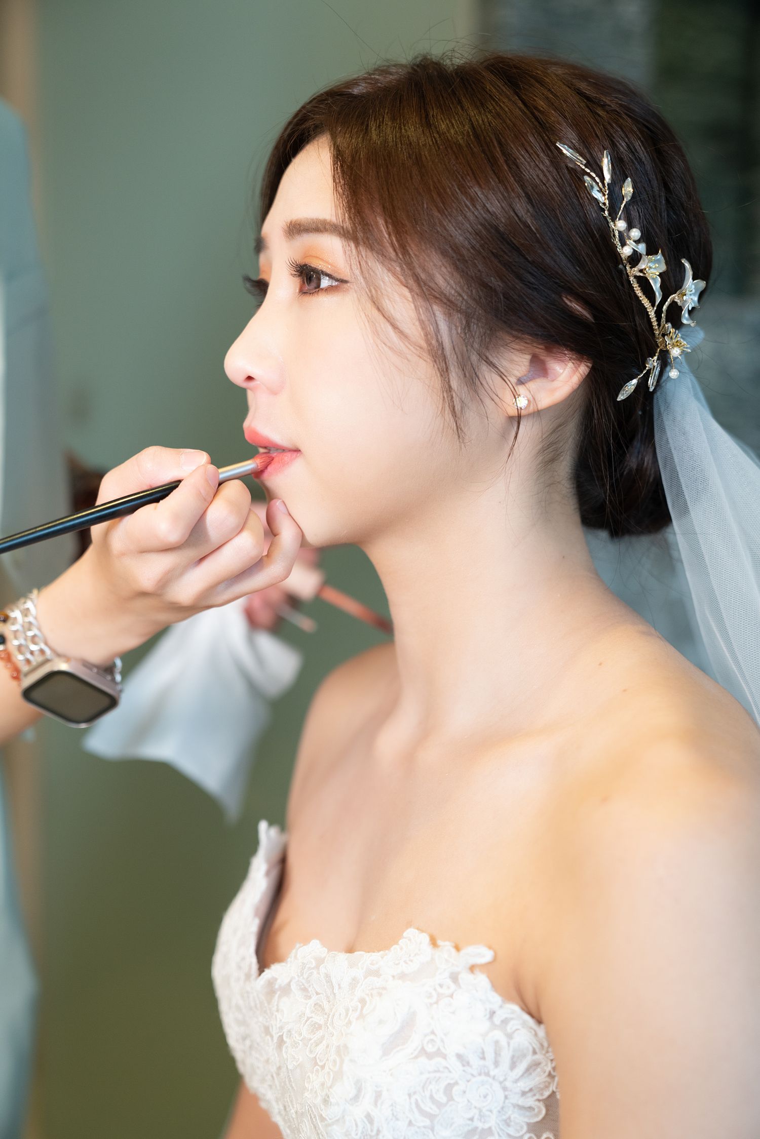 A bride in a white lace wedding dress getting makeup applied while wearing a delicate silver hair accessory and veil.