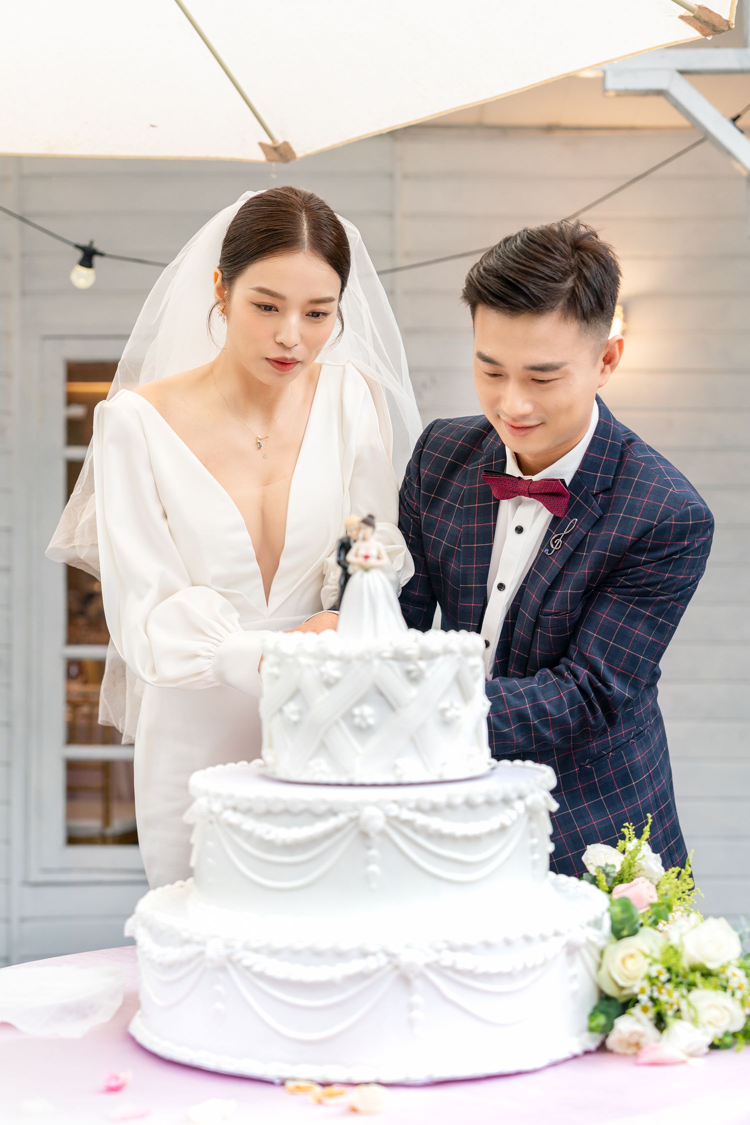 A wedding couple stands together at their reception looking down at their elegant three-tiered white wedding cake.