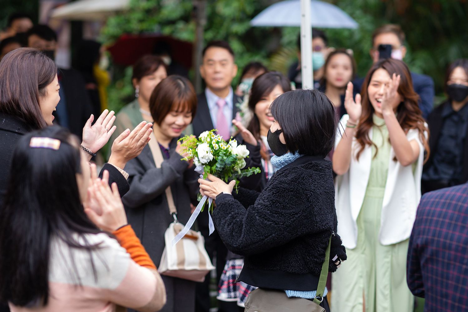 A crowd gathers outdoors for a celebratory moment with bouquets and clapping at a festive event.
