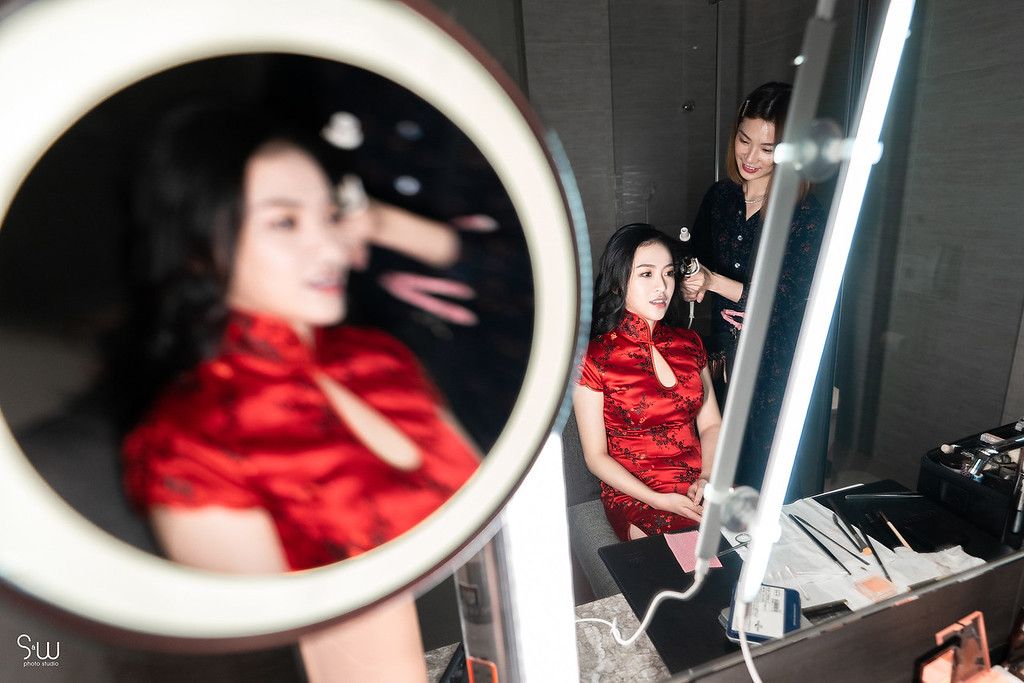 Reflection in makeup mirror shows bride getting ready in red Chinese dress.