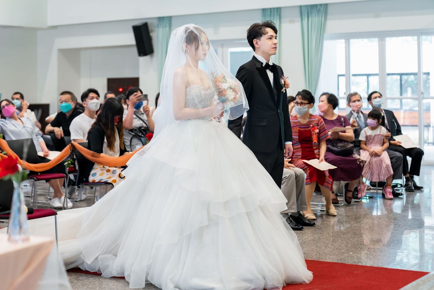 Wedding ceremony scene with couple standing before seated guests in bright venue.