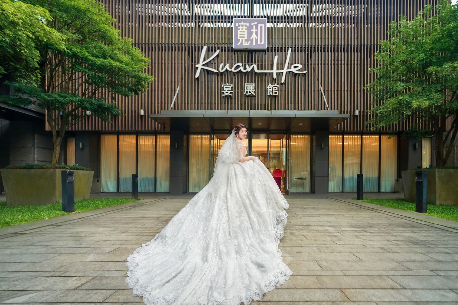 A flowing white wedding gown with a long train is displayed in front of the Kuan Life building entrance.