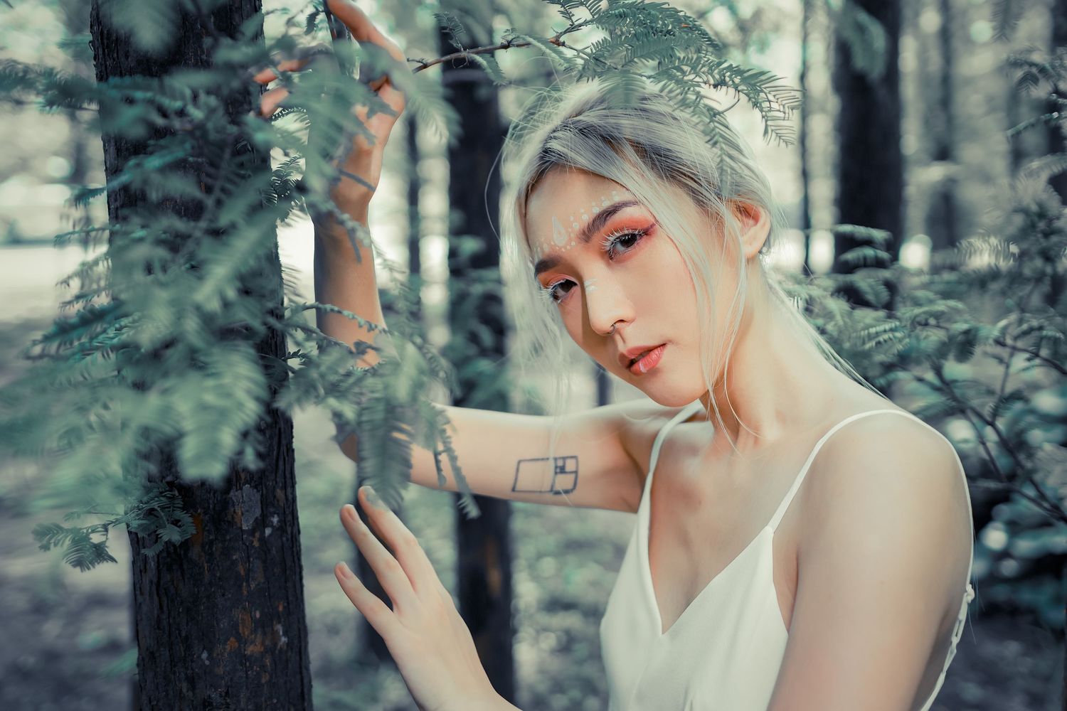 Artistic portrait in a forest setting with dreamy teal color tones and delicate white camisole among tree branches.