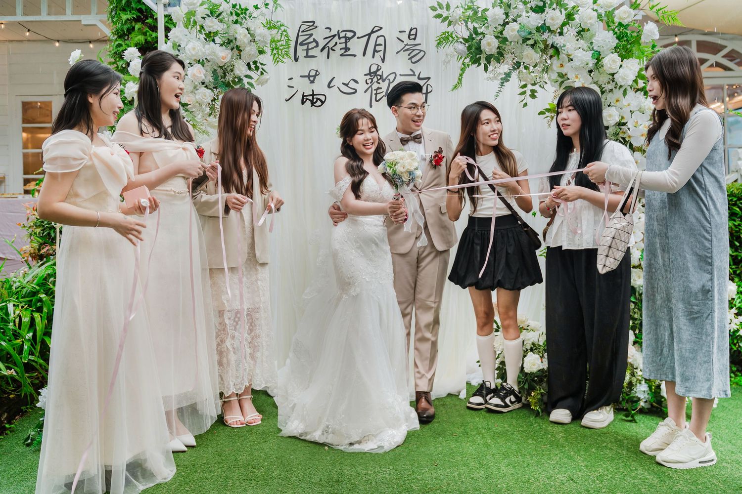 Wedding party gathering outdoors in front of floral arch with guests in coordinated pink and beige dresses during celebration.