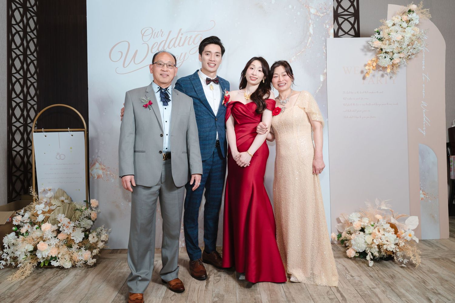 Elegant wedding party group pose in formal attire against a grey backdrop with white floral decorations.