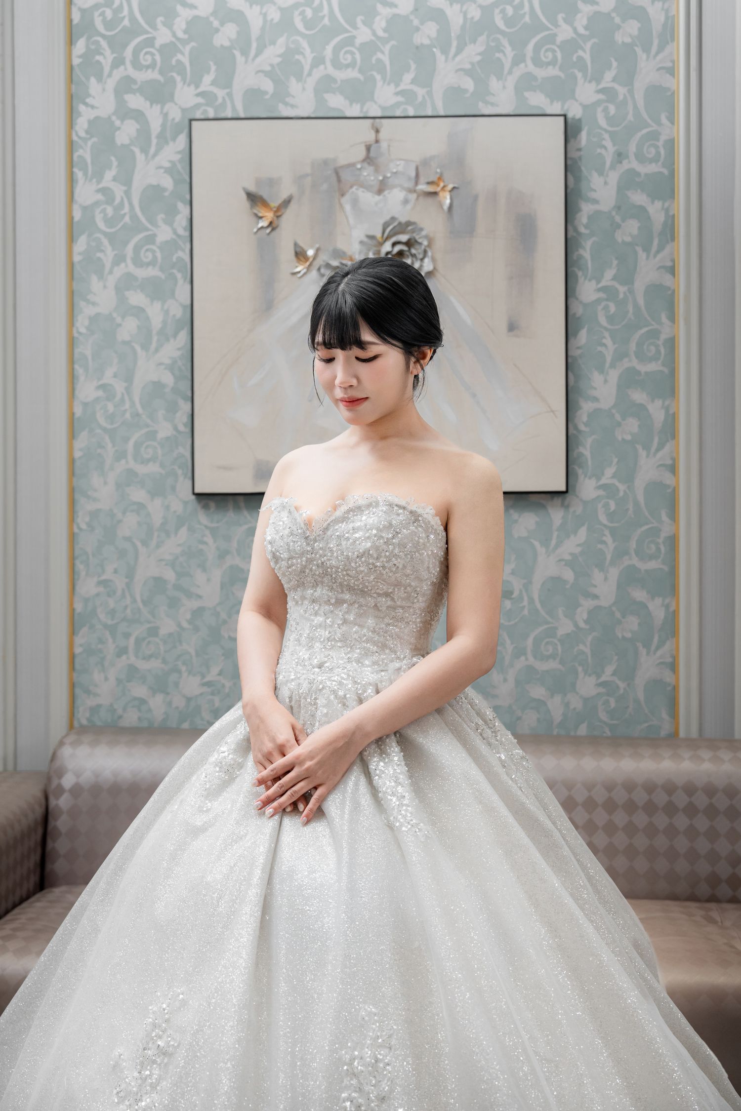A bride in a strapless white ballgown with intricate beading poses gracefully in an elegant room with floral wallpaper.