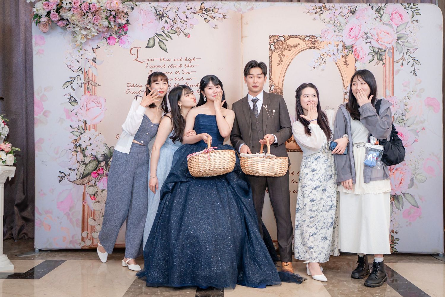 Group of five people posing at a floral wedding backdrop, woman in blue gown holding wicker baskets.