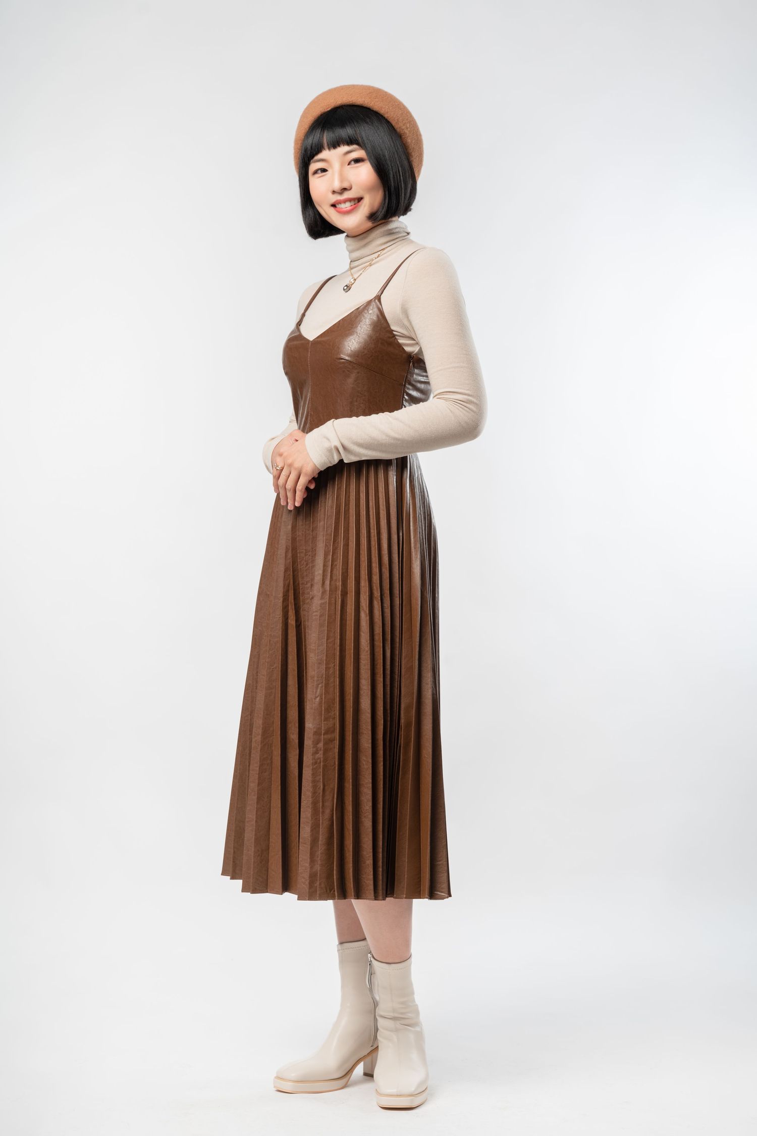A person poses in a brown corduroy pinafore dress layered over a beige turtleneck sweater with matching boots.