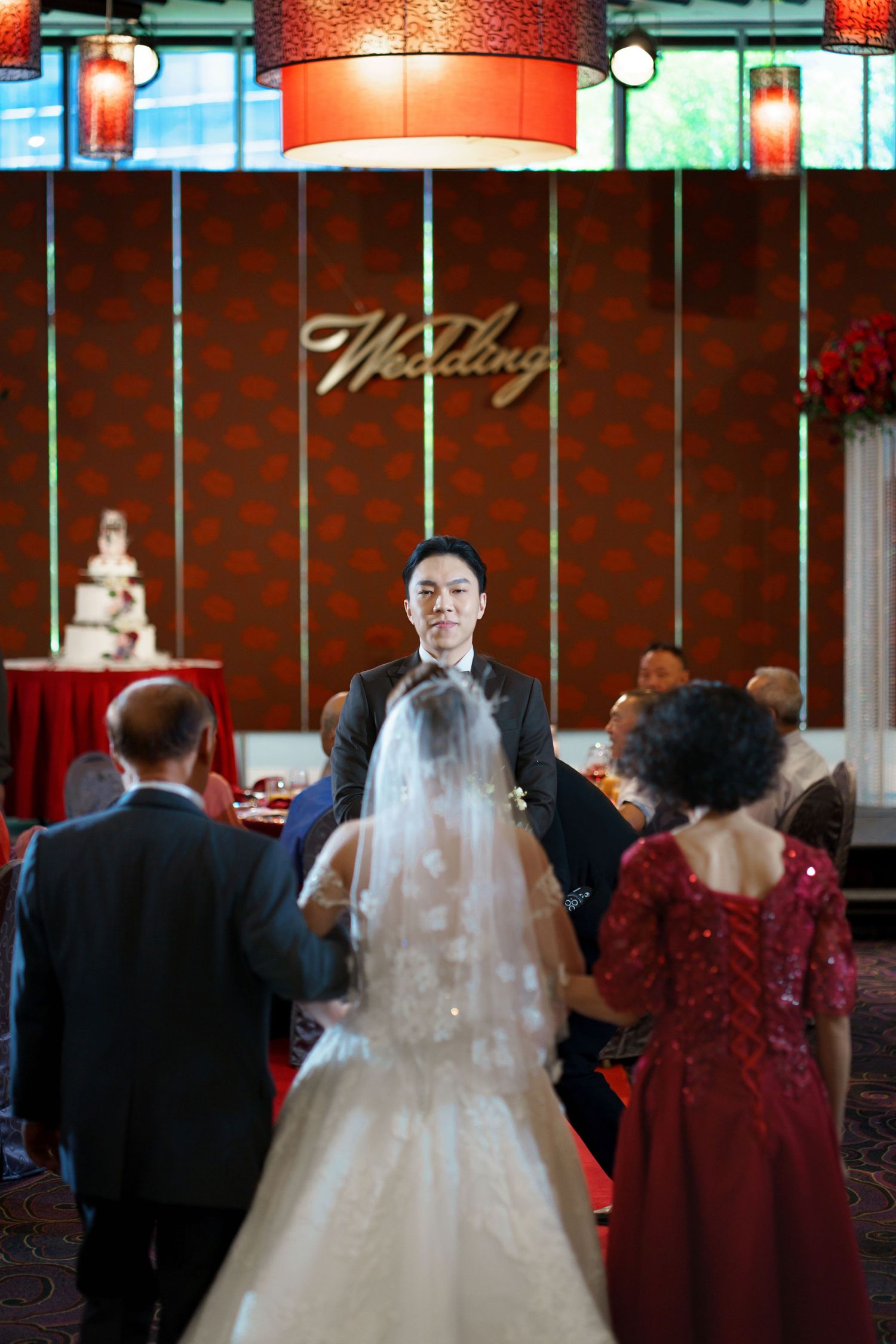 Wedding party in burgundy bridesmaid dresses and formal attire gather during indoor reception with warm lighting.