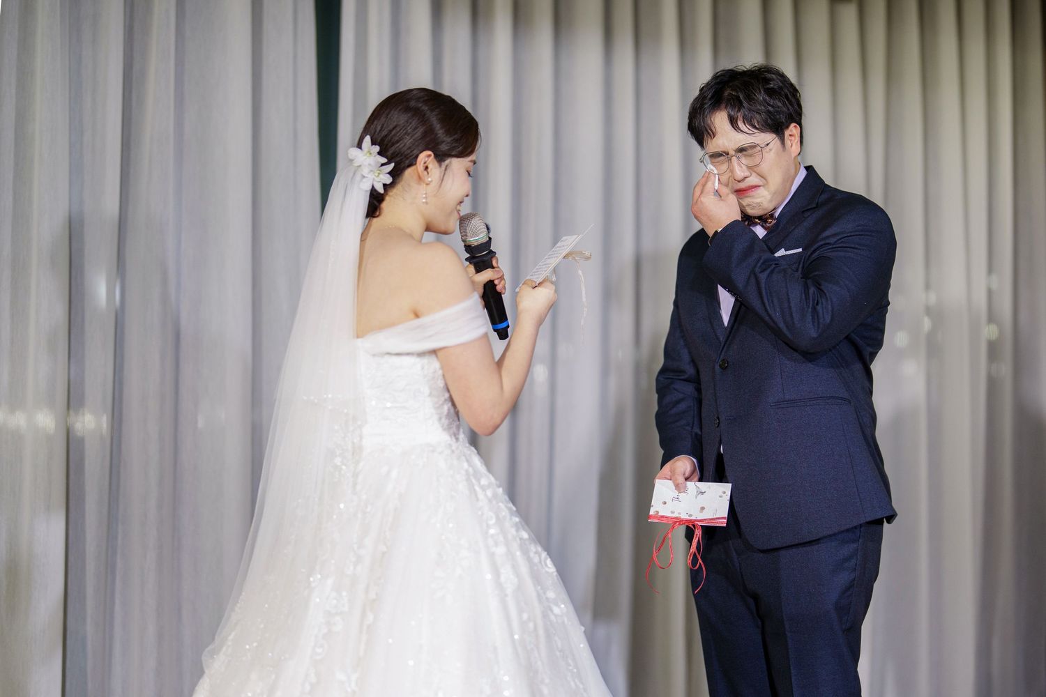 A bride in a white lace gown and groom in navy suit share an emotional moment during their wedding ceremony.