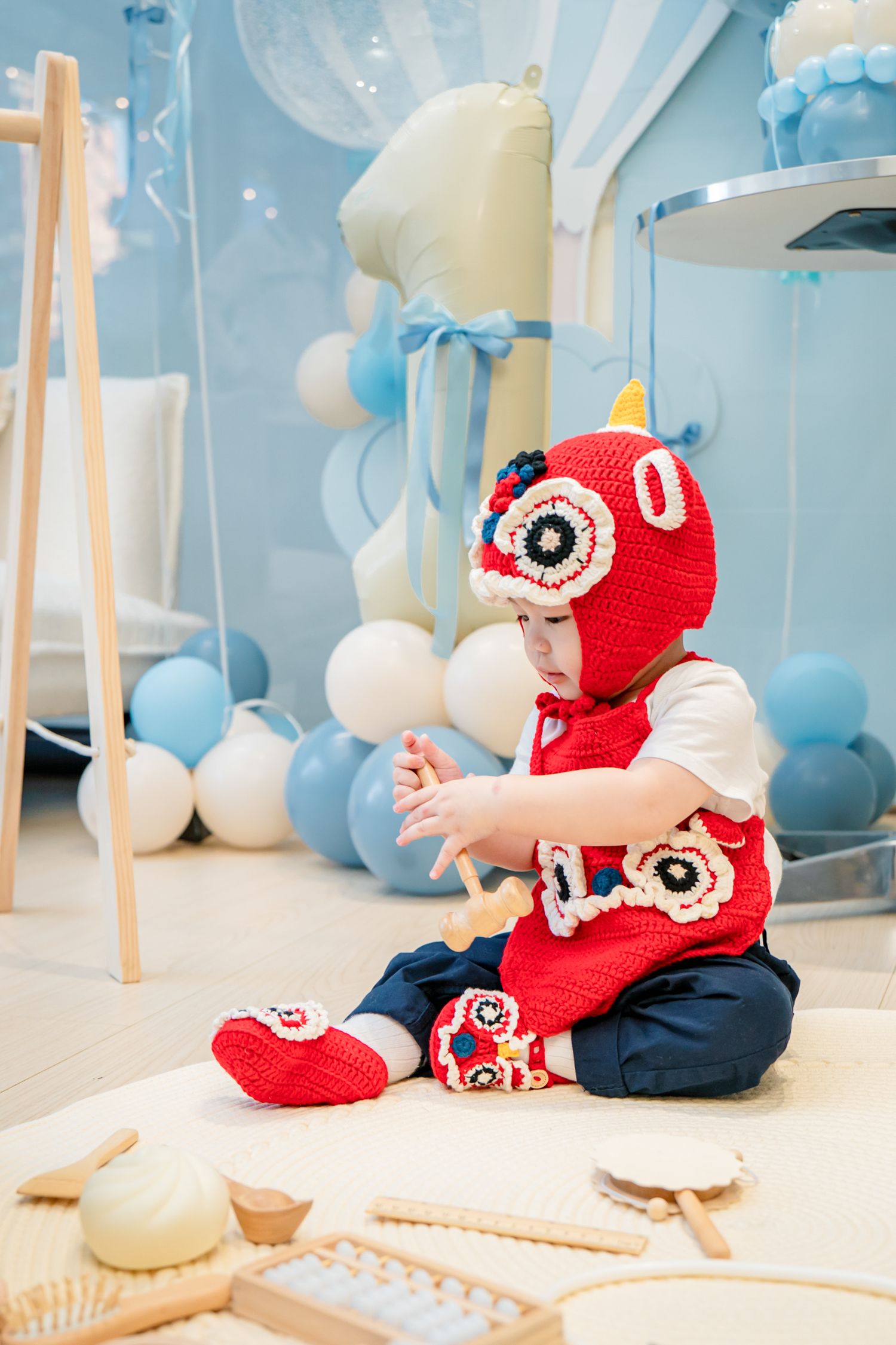 Baby wearing red crochet owl hat sits on white blanket with blue balloon decorations in background.