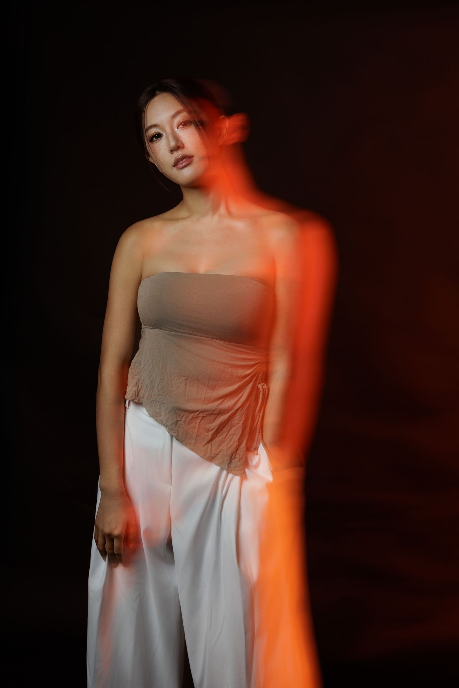 A woman in a strapless beige top and white wide-leg pants poses dramatically under warm red lighting against a dark background.