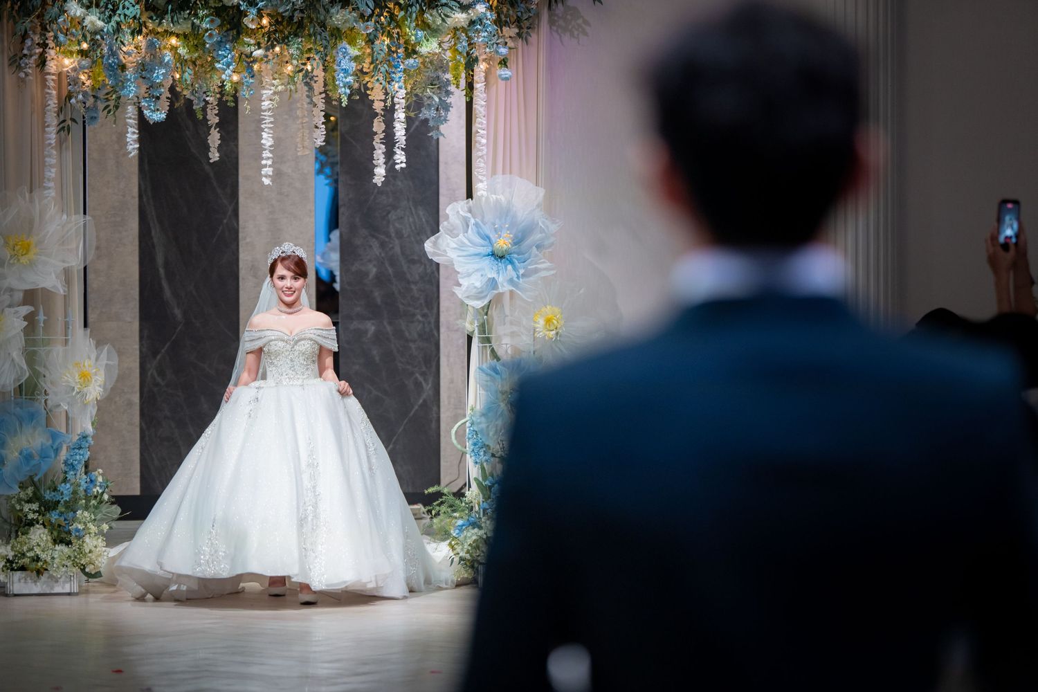 Bride in white ball gown wedding dress stands before groom at elegant floral-decorated wedding venue.