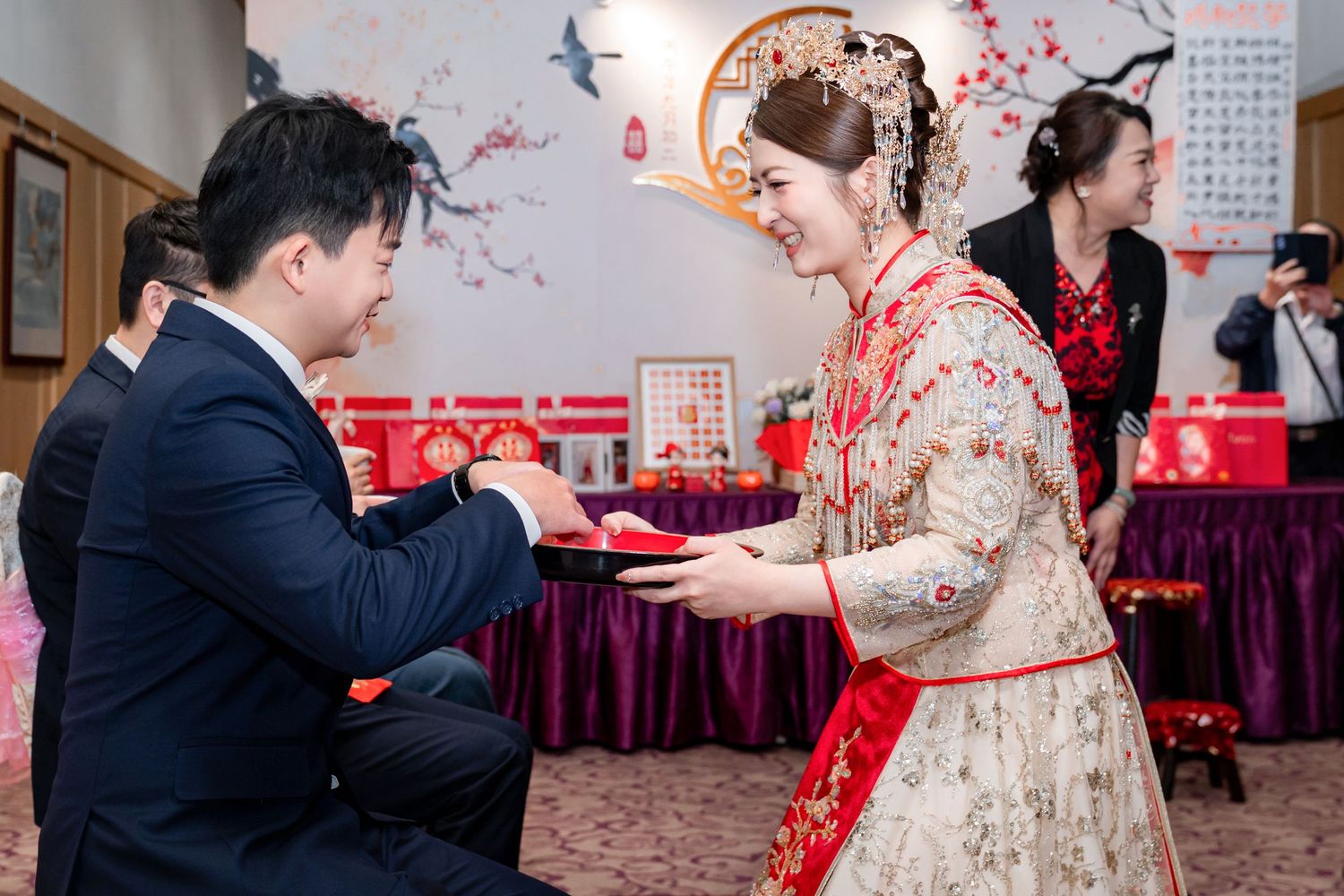 Bride in an ornate red and gold traditional Chinese wedding gown offers a tray to a kneeling groom during a tea ceremony.