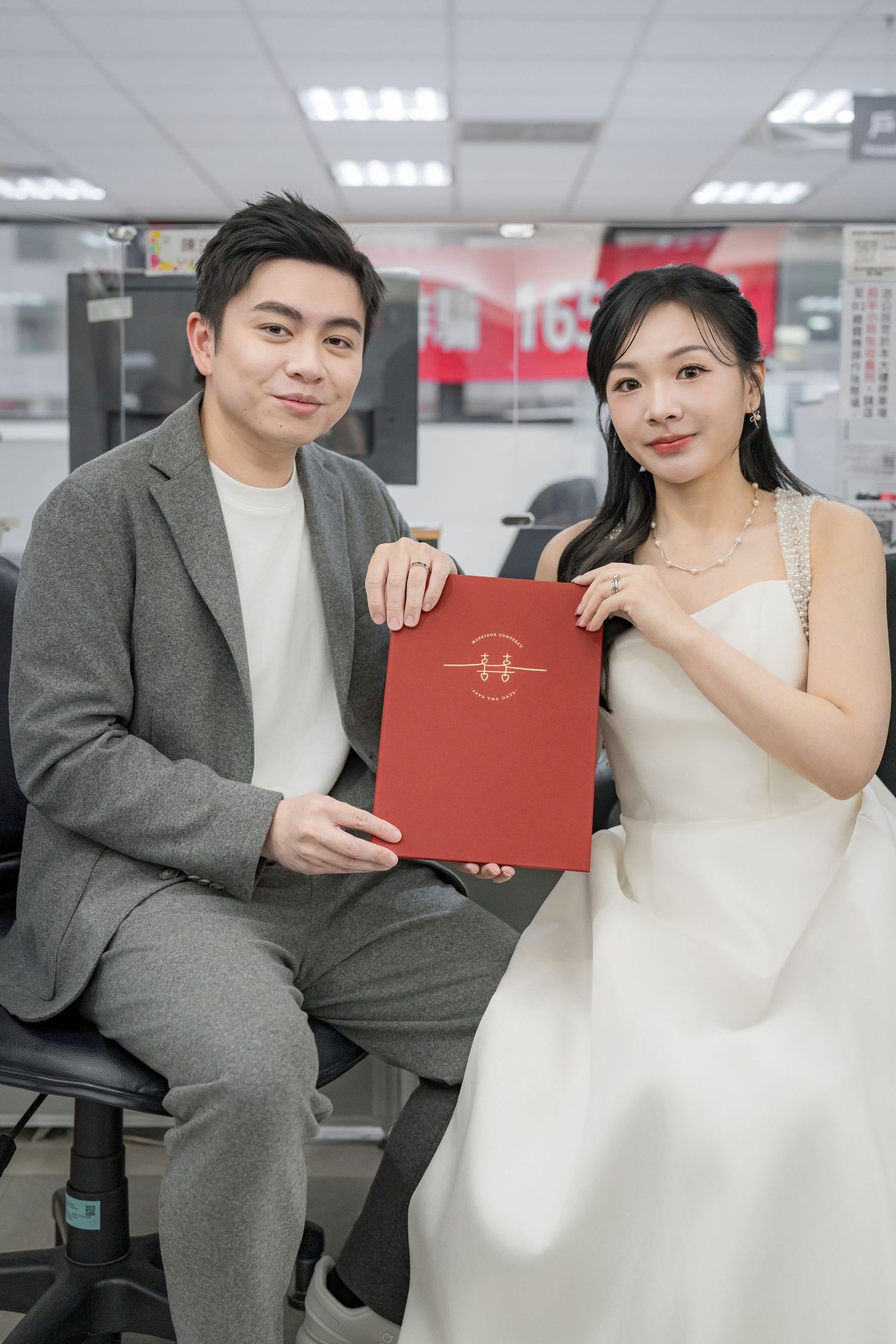 A couple in formal attire poses together holding a red document folder in an office setting.