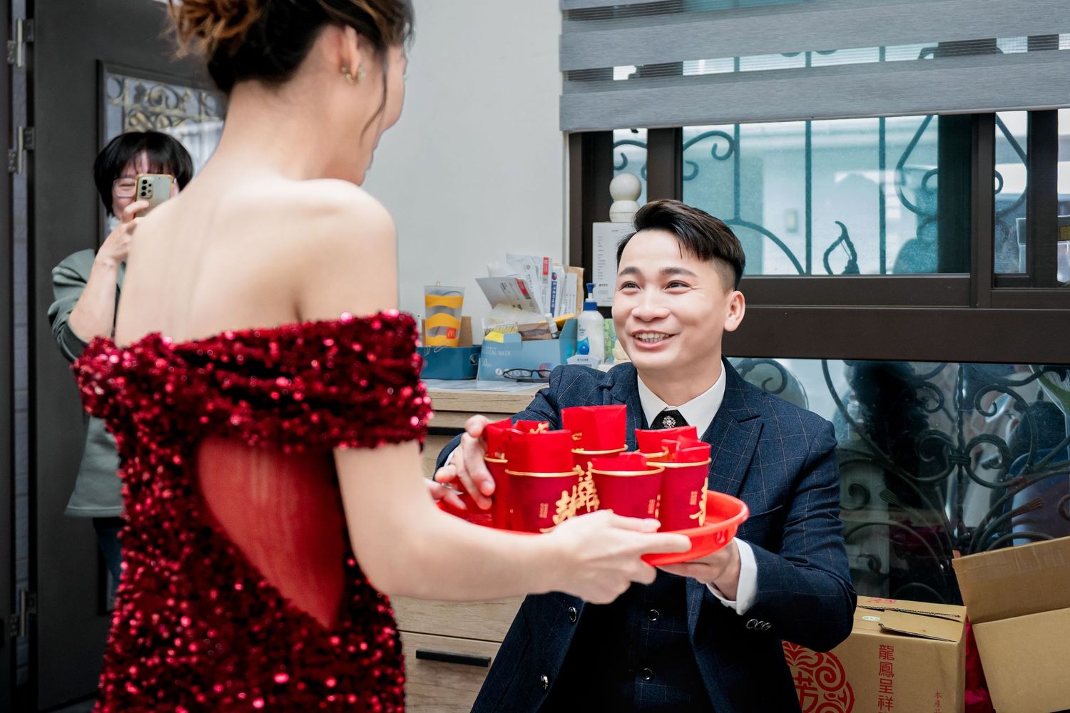 A group of people in formal attire gather around a table with red decorations and boxes at a holiday event.