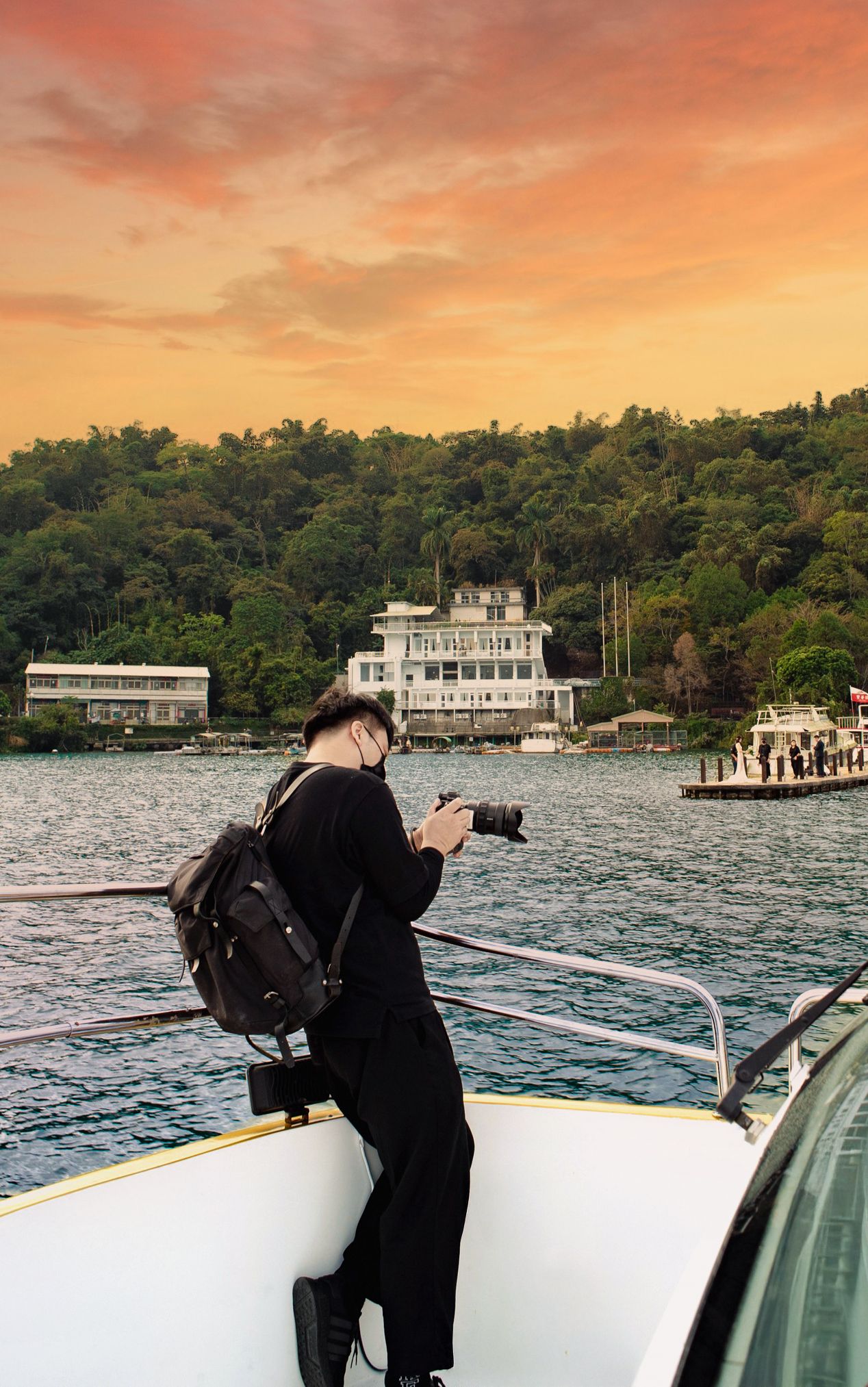 Photographer on a boat captures waterfront mansion at sunset with orange sky and lush green trees in background.