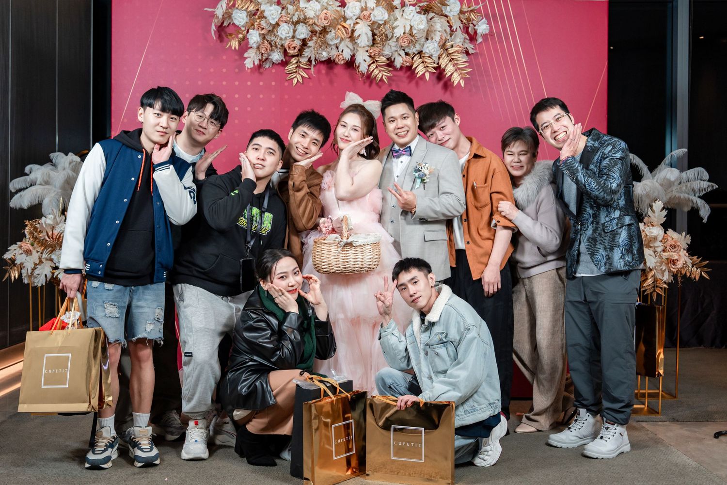 Festive gathering with people posing against pink background with hanging white floral arrangements.