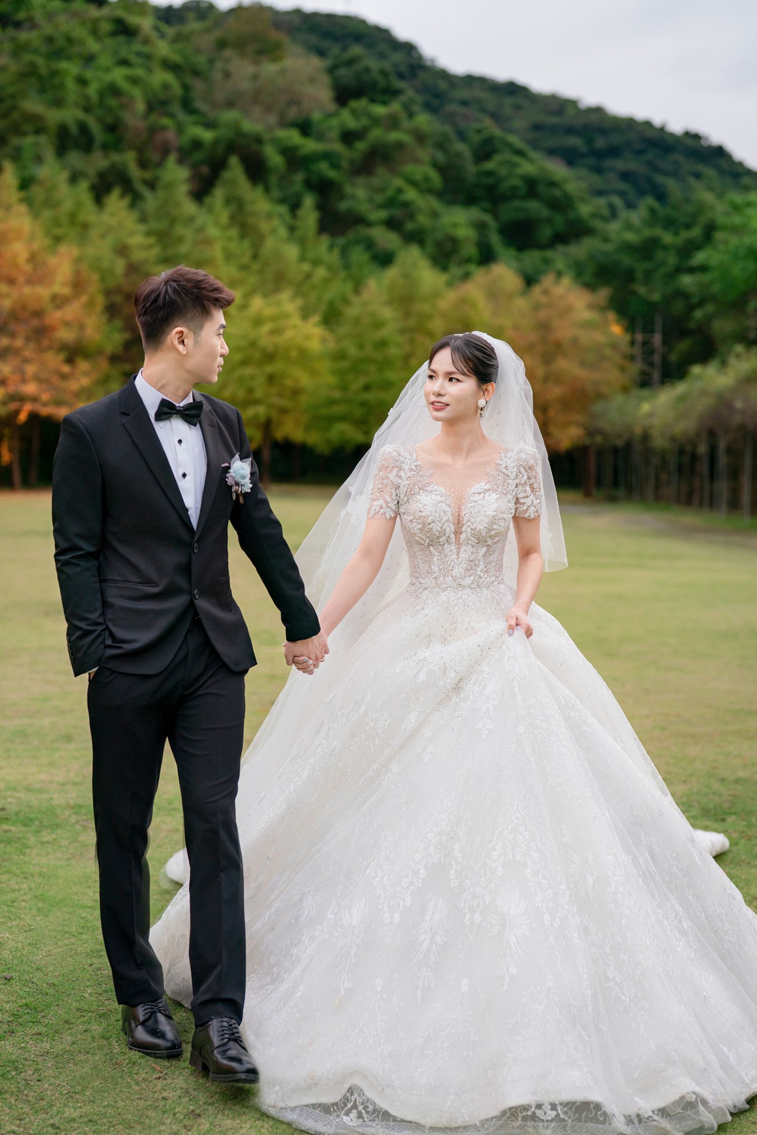 A romantic outdoor wedding photoshoot with a couple in formal attire walking through a grassy field near wooded hills.