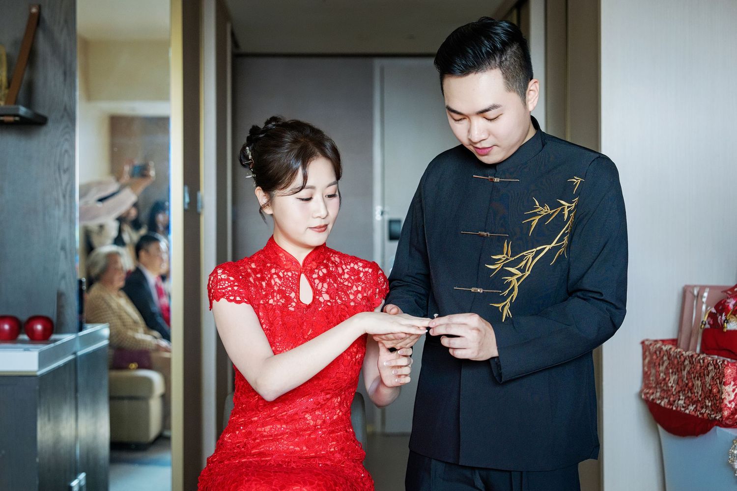 Couple in traditional Chinese wedding attire exchanging items during tea ceremony in a home setting.