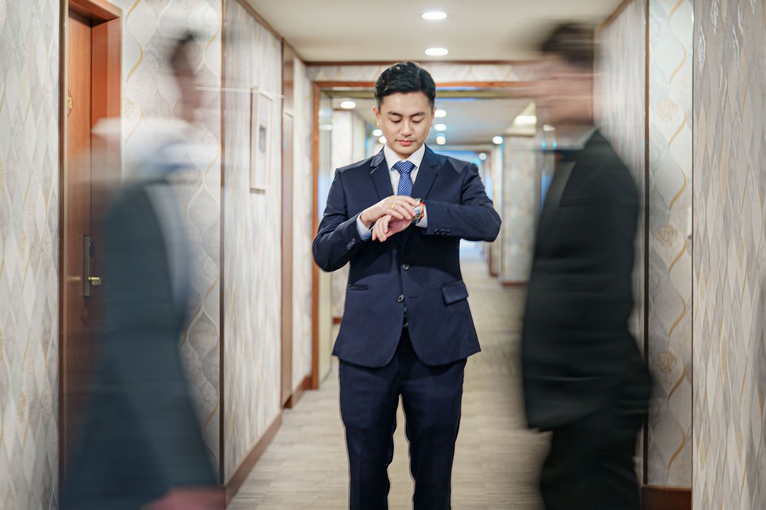 Businessman in navy suit checks smartphone while walking down a modern office hallway with wood paneled walls.