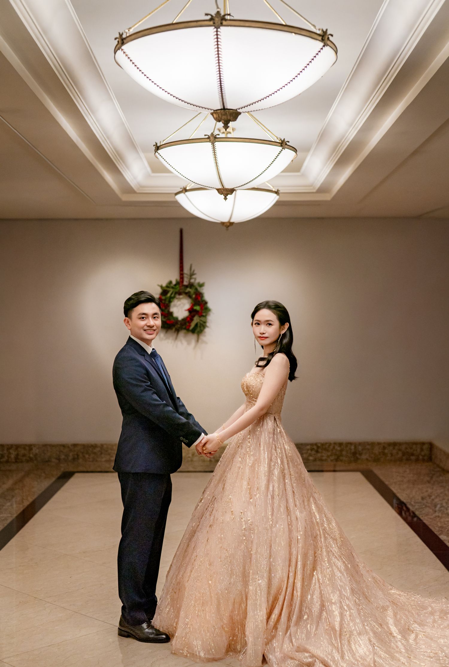 A couple in formal attire dances in an elegant ballroom with ornate lighting fixtures overhead.
