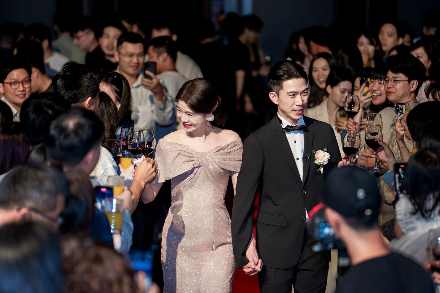 Couple walks through crowd on red carpet at formal event as spectators take photos with phones.