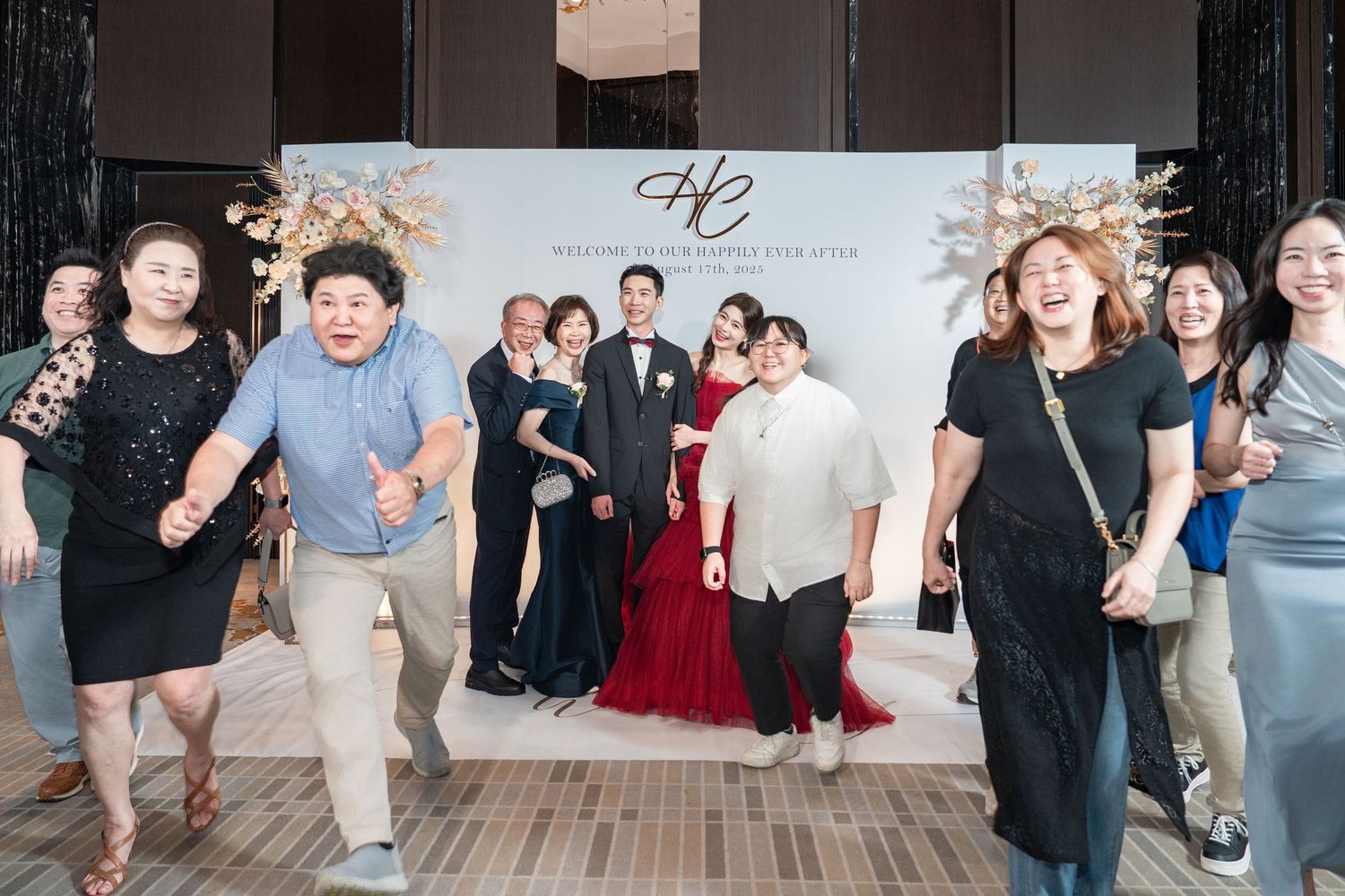 Wedding guests dance and celebrate in front of a white backdrop at an indoor reception venue.