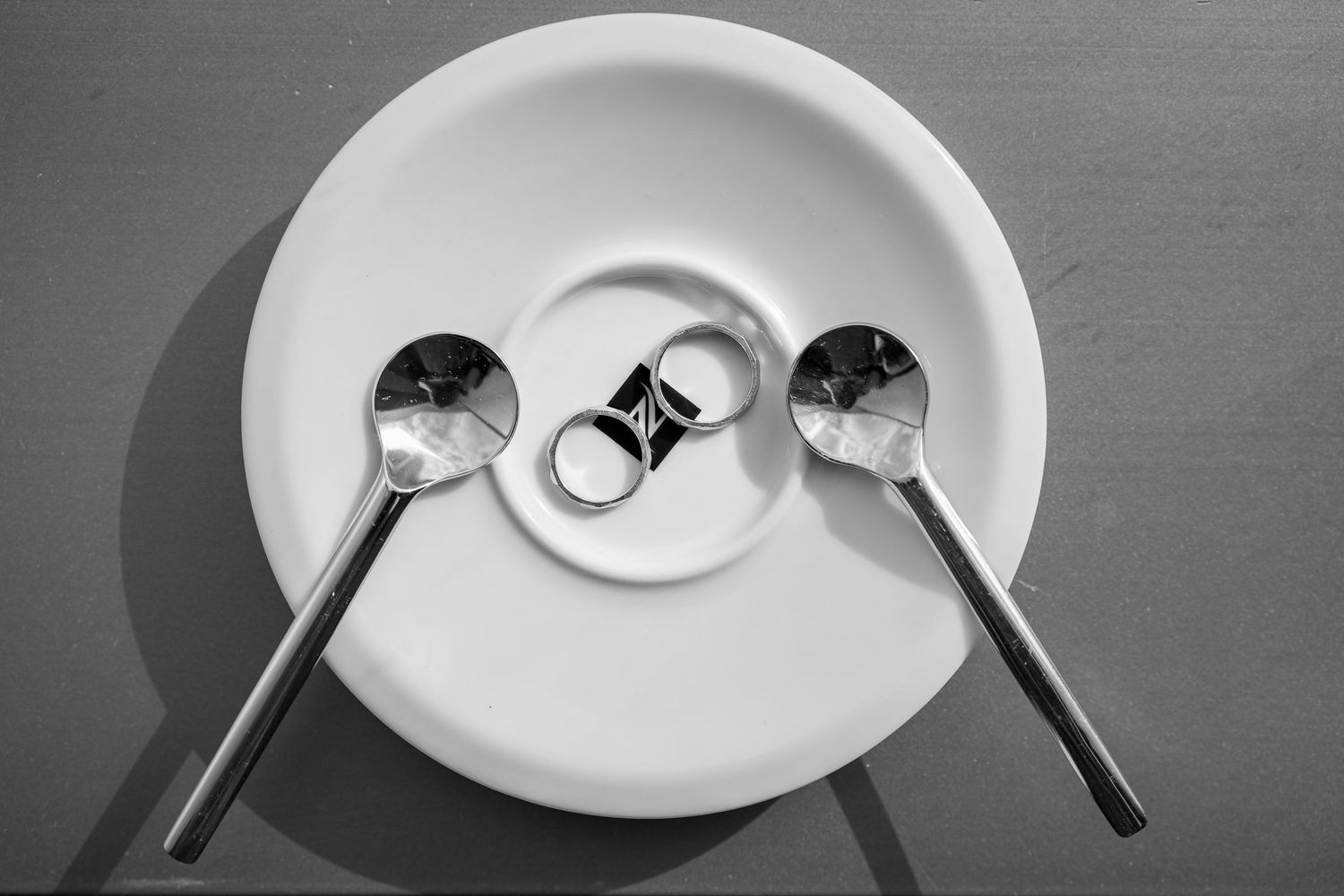 A minimal black and white photograph of two spoons and a ring arranged symmetrically on a white plate.
