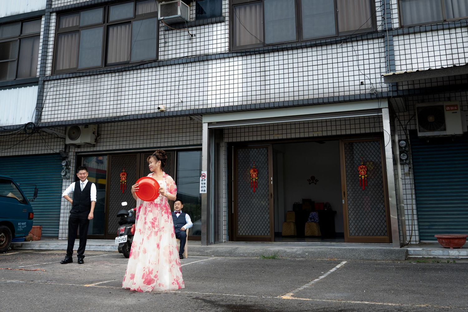 Woman in traditional Chinese wedding dress holding red fan outside urban building with garage doors.
