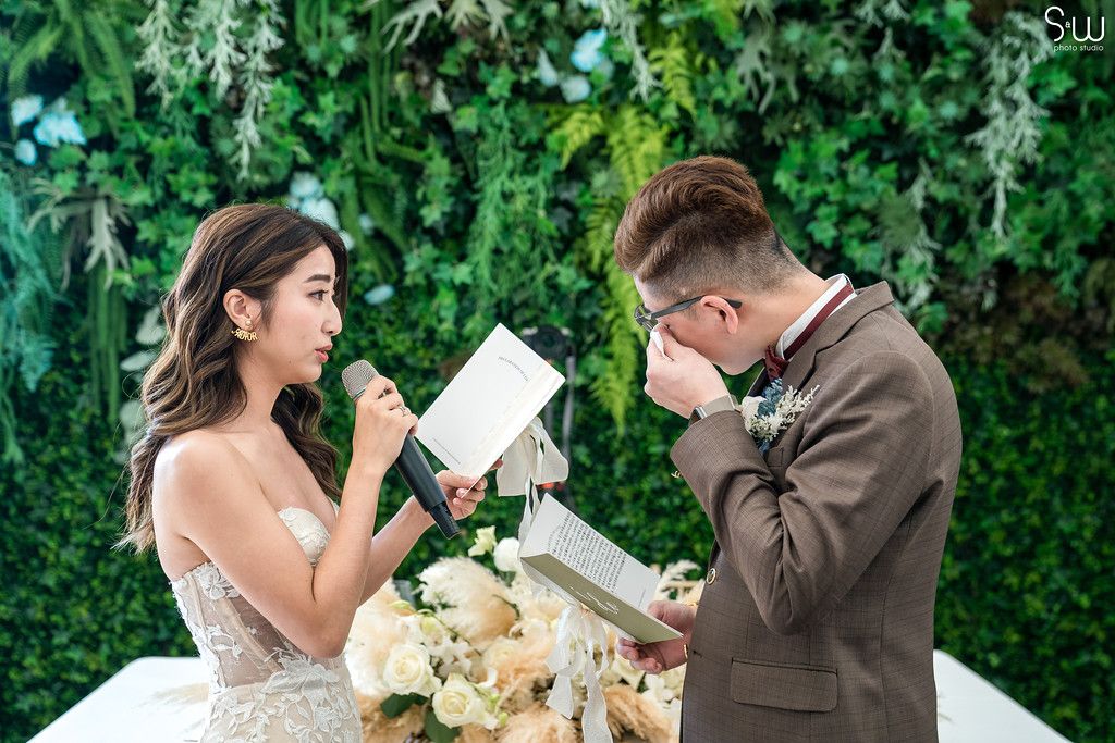 Emotional wedding ceremony moment with couple reading personal vows against green botanical backdrop.