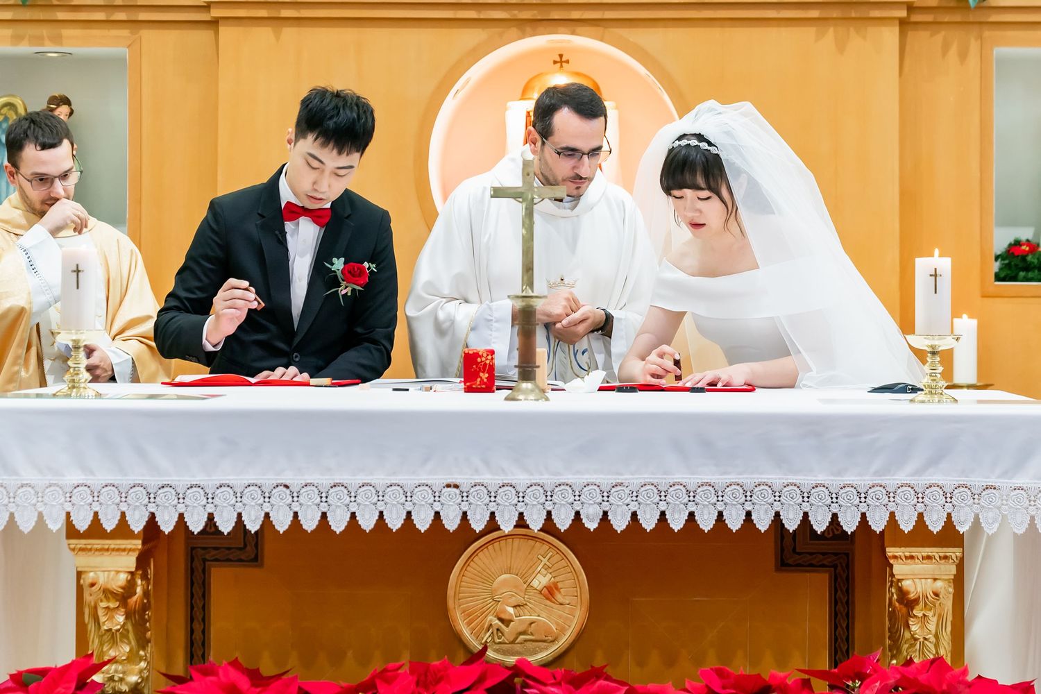 Wedding ceremony participants signing documents at decorated altar table during Catholic mass.