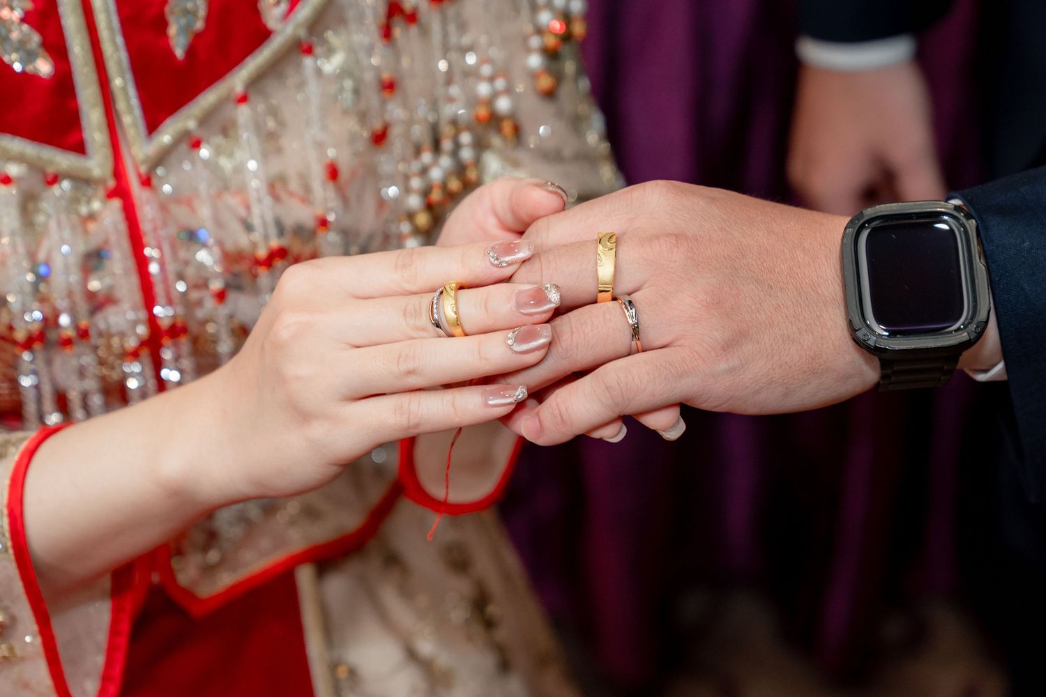 Close-up of a bride in a red and gold embroidered outfit exchanging rings with a groom wearing a smartwatch during a wedding ceremony.