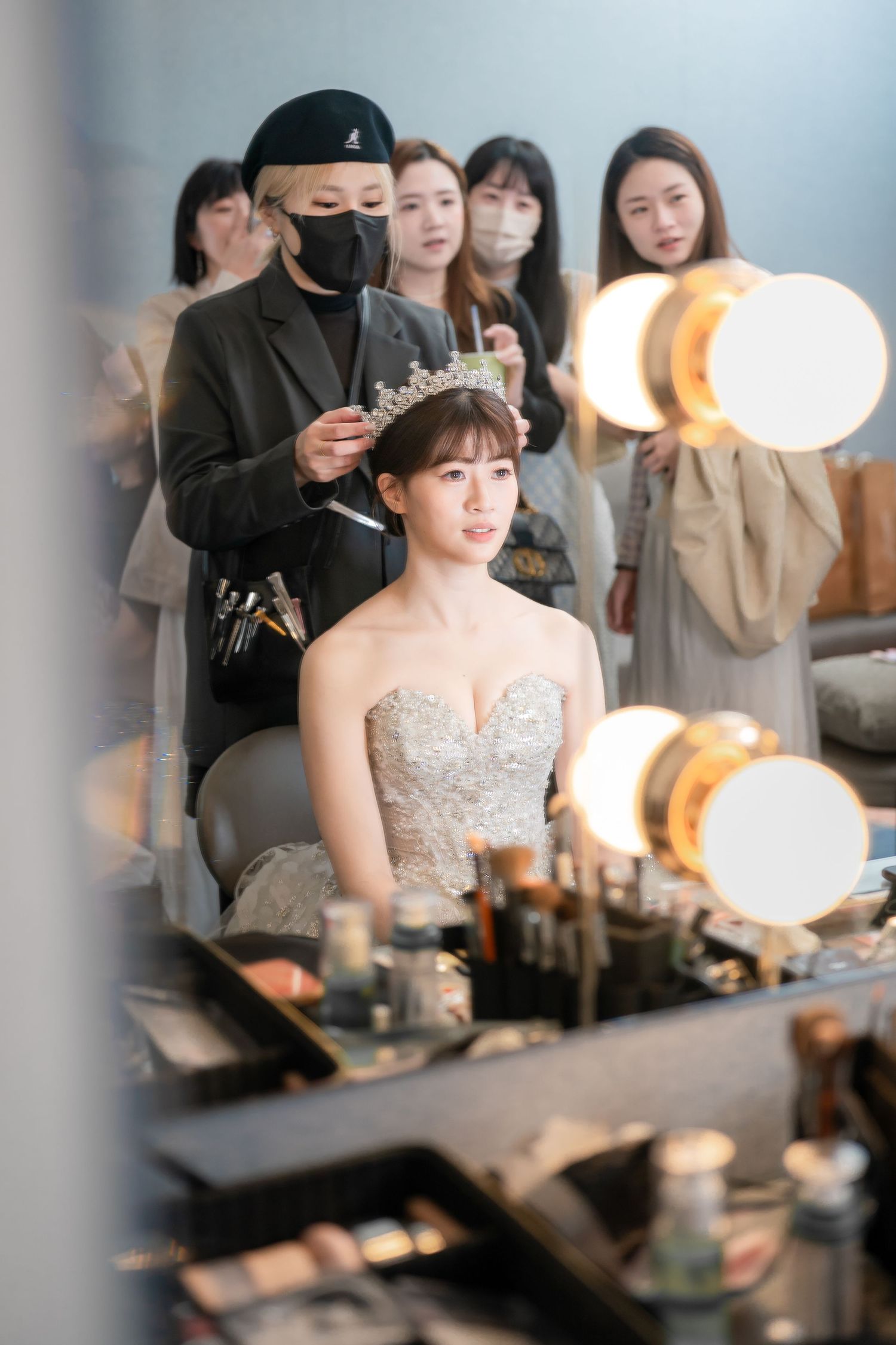 Bride in strapless gown and tiara gets hair styled by makeup artist at vanity mirror surrounded by guests.