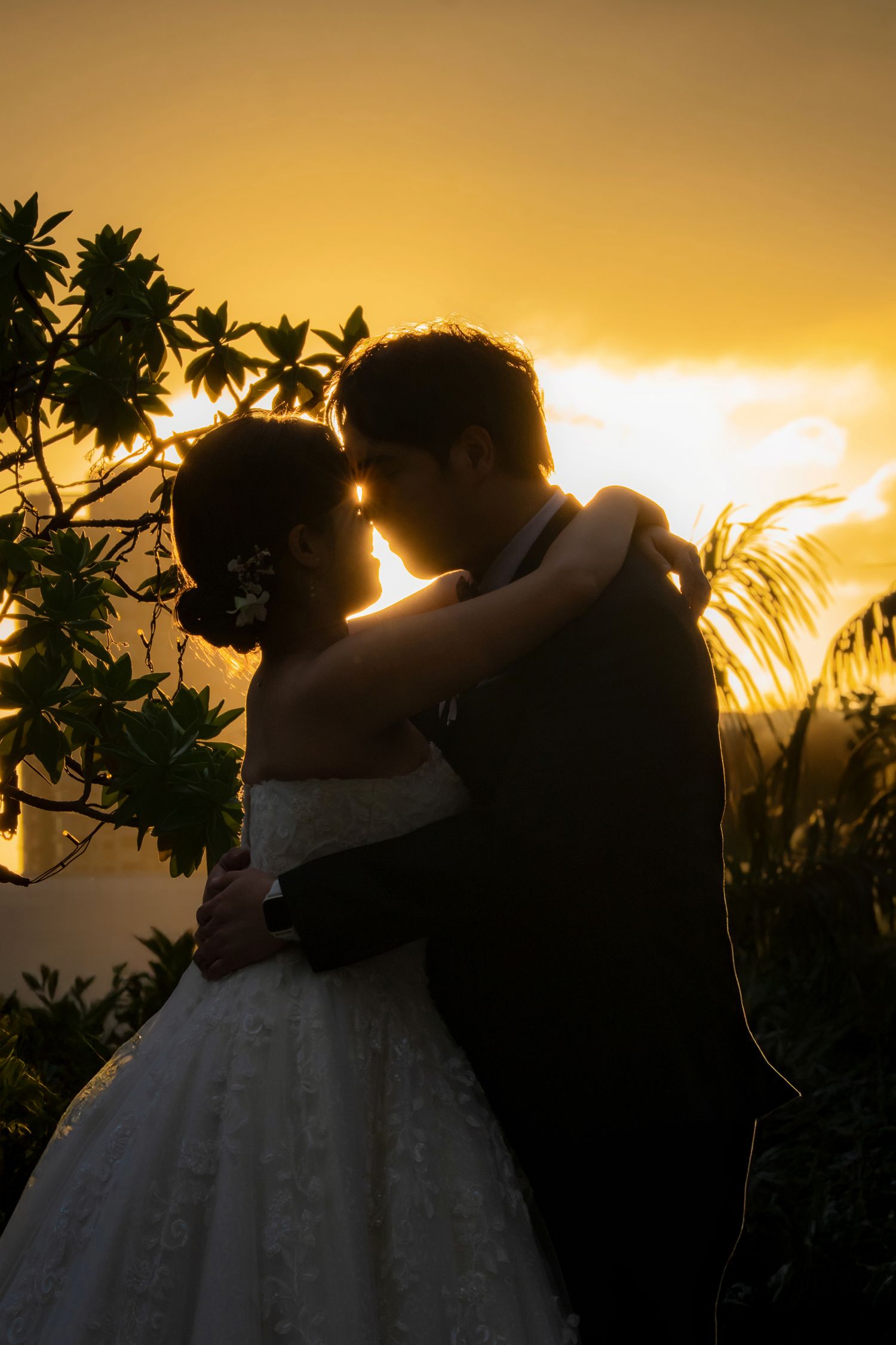 A romantic silhouette of a couple in wedding attire embracing at sunset with palm trees and golden sky in the background.
