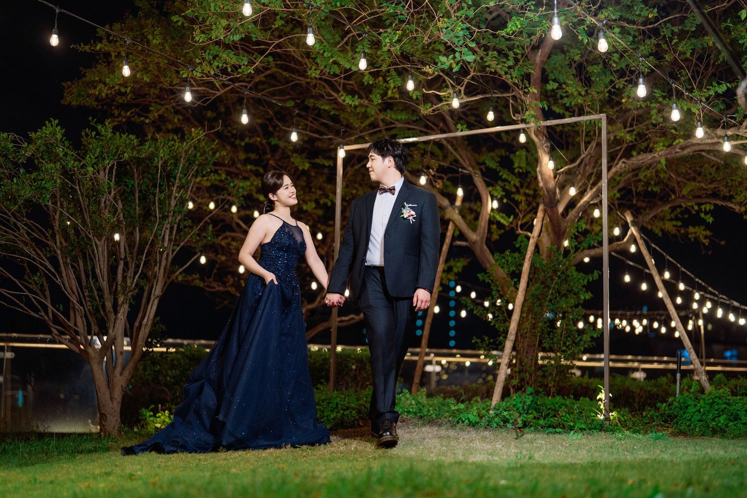 String lights illuminate an outdoor evening wedding reception area with elegant tables and natural garden surroundings.