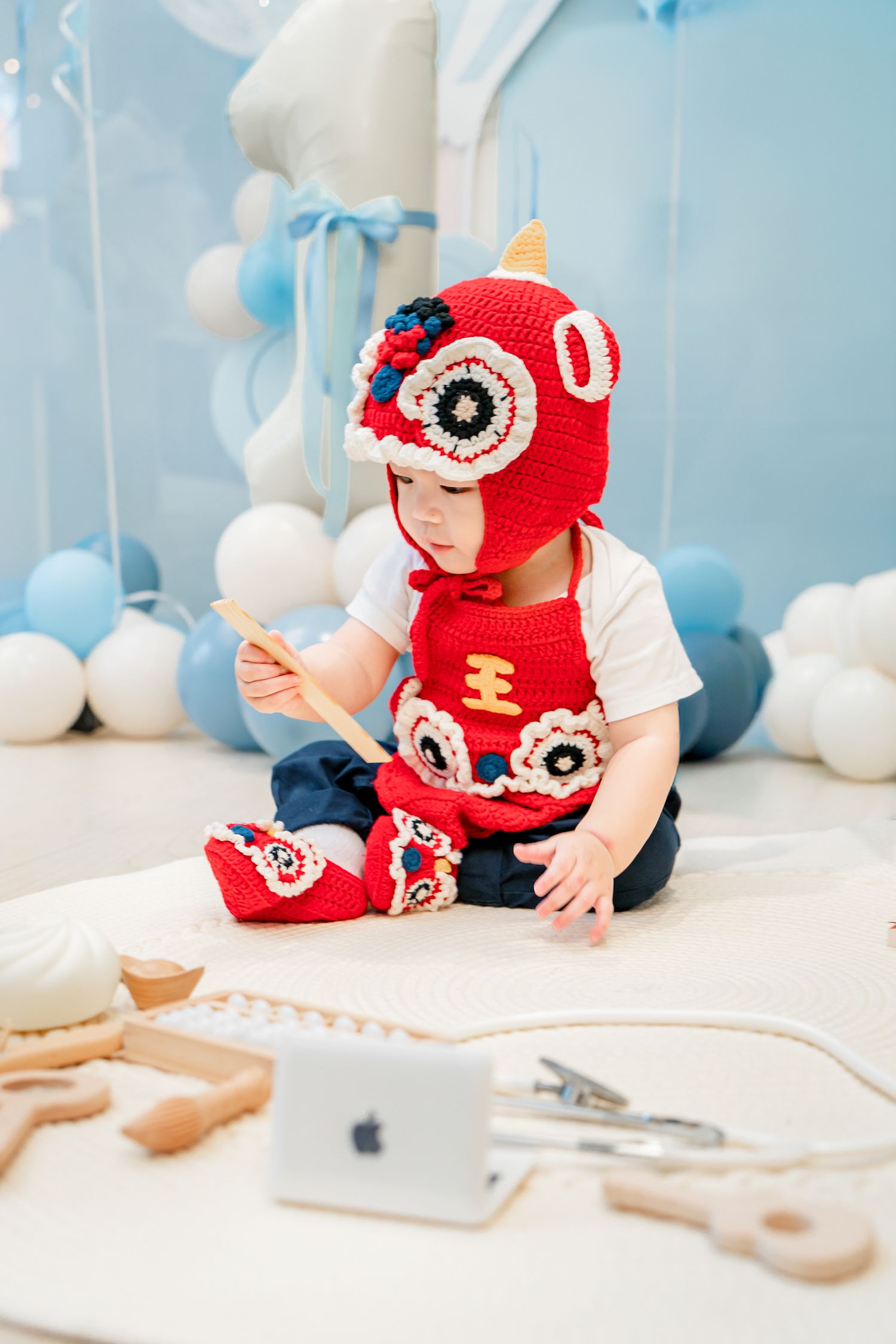 Baby wearing red crochet hat plays with tablet device on white floor with blue balloons in background.