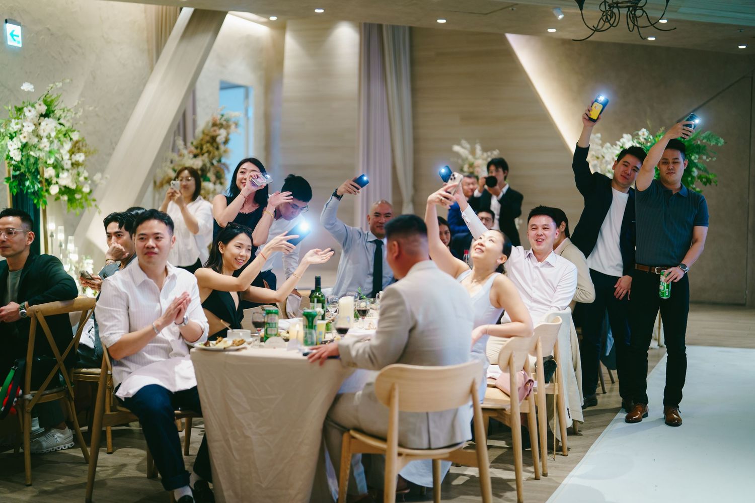 Guests celebrate at a modern wedding reception in a bright banquet hall featuring long wooden tables and decor.
