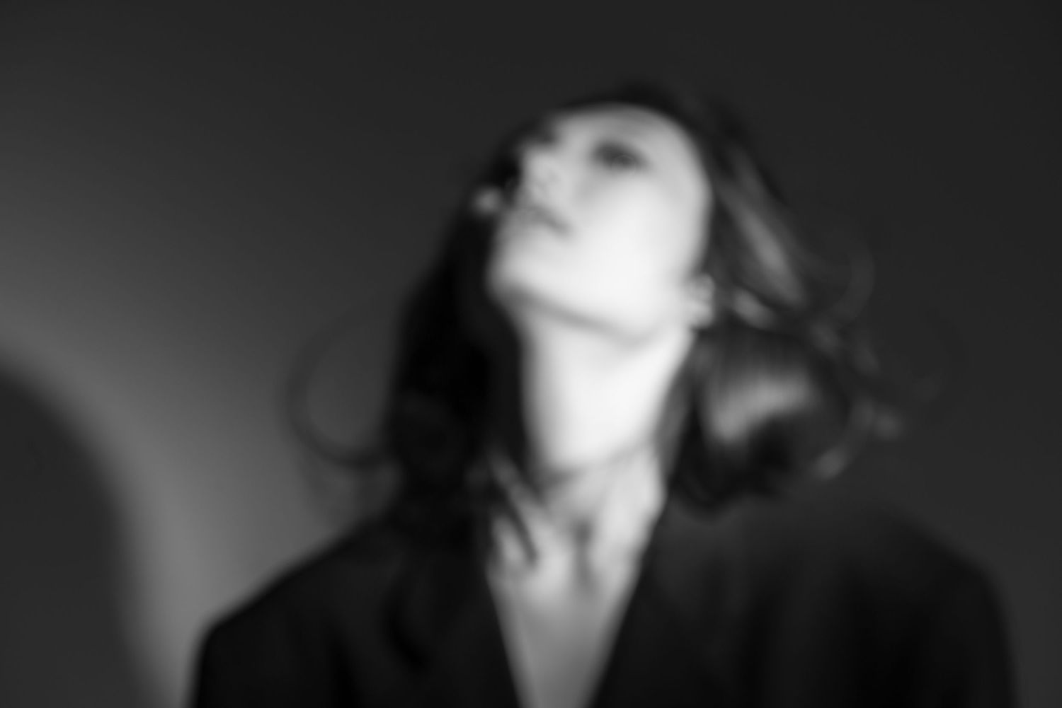 Artistic black and white blurred portrait of a figure tilting their head back with flowing hair against a dark background.