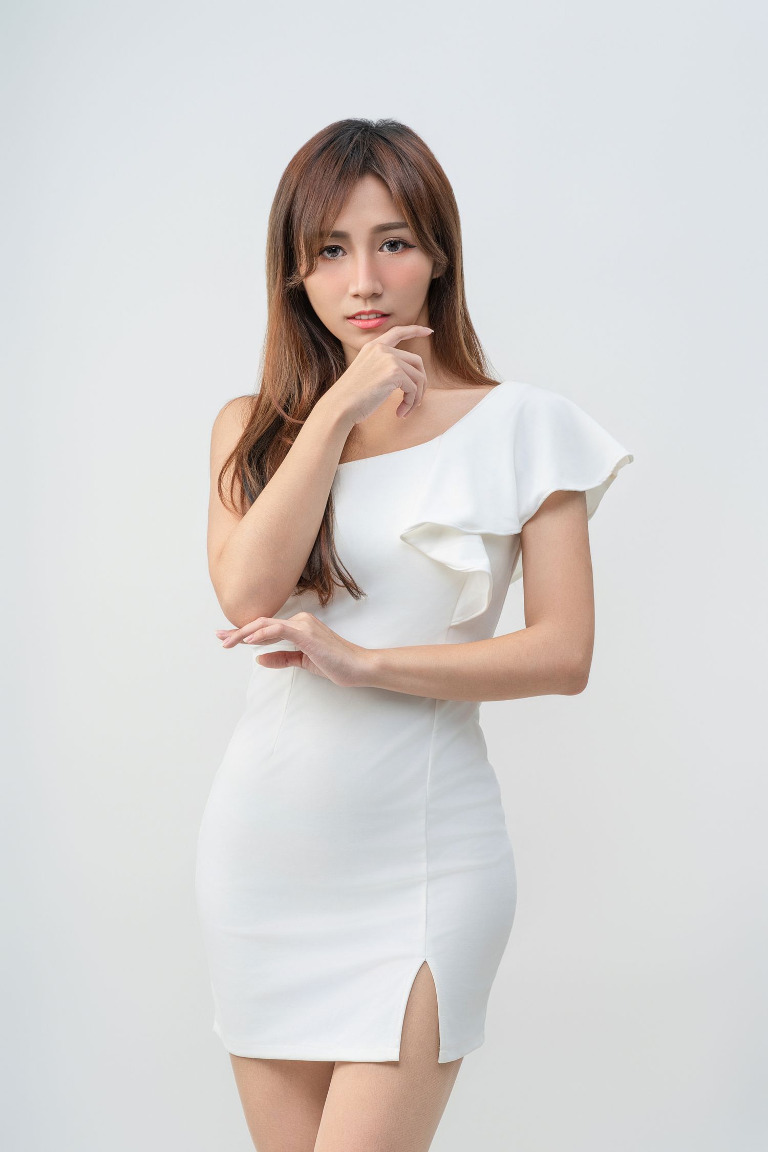 Elegant woman in white one-shoulder bodycon dress with ruffle detail and side slit poses pensively against light background.