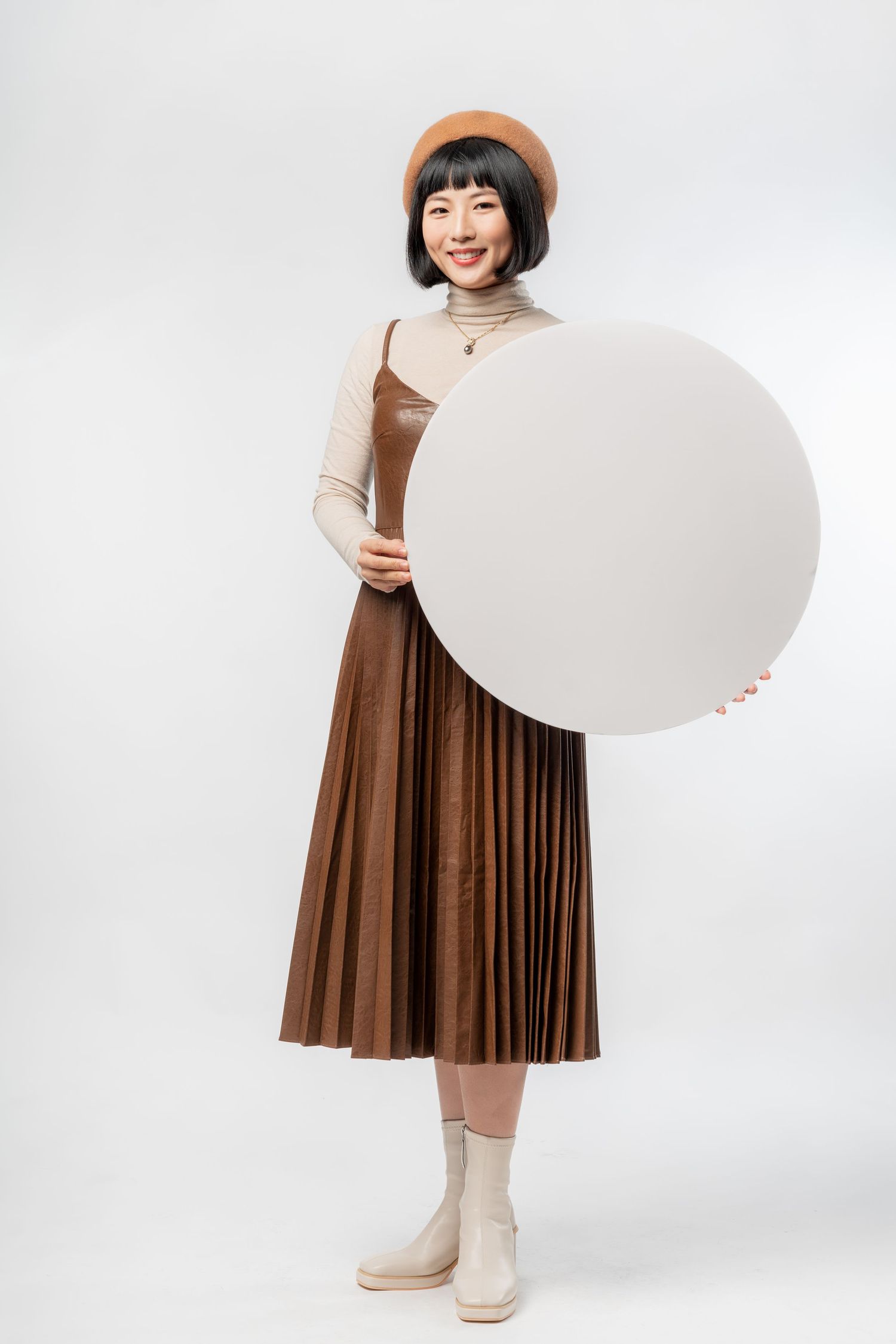 Stylish person in brown pleated dress and beret holding large white circular blank sign against white background.