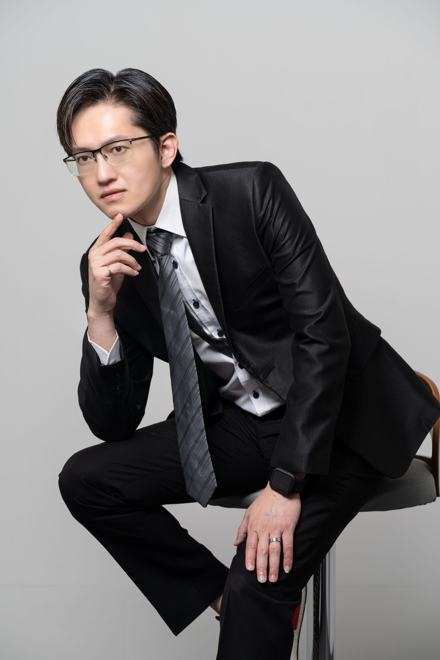 Professional portrait of figure in black suit and tie wearing glasses sitting on stool in contemplative pose against gray backdrop.