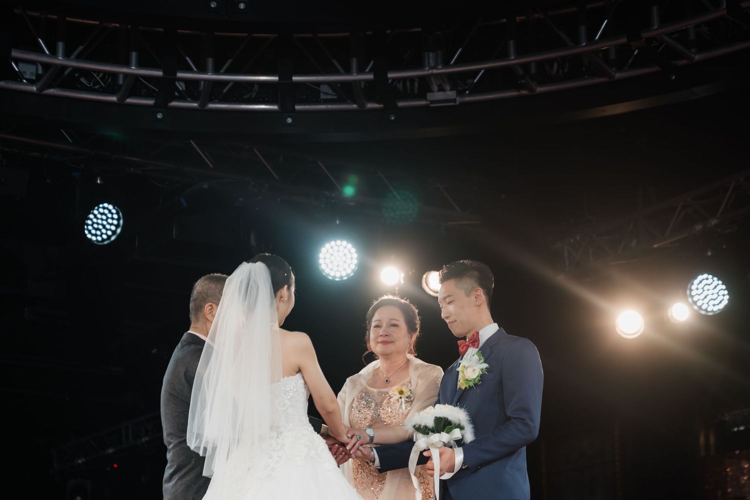 A dark indoor wedding ceremony scene with spotlights illuminating a couple and officiant during their exchange of vows.