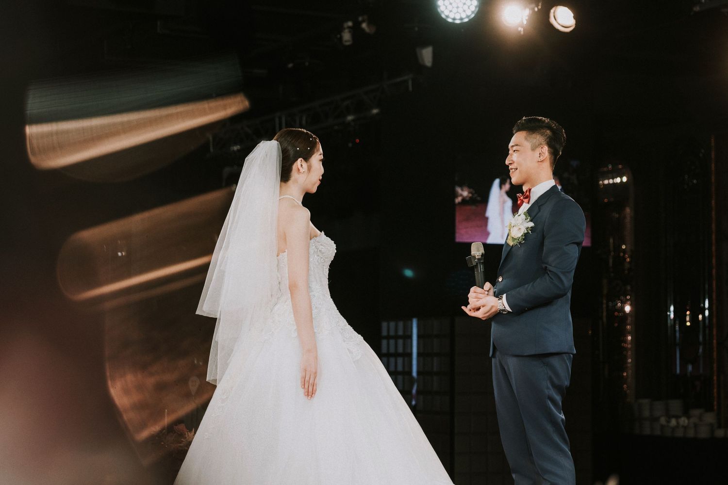 A couple stands facing each other during their wedding ceremony at a dimly lit indoor venue with spotlights glowing above.