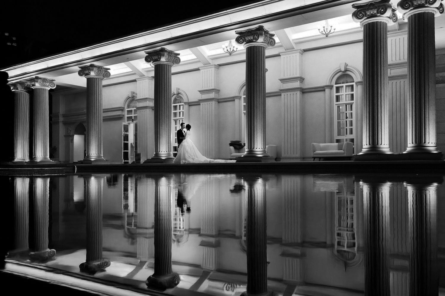 A dramatic nighttime reflection of neoclassical columns and architecture in a still pool of water, captured in black and white.