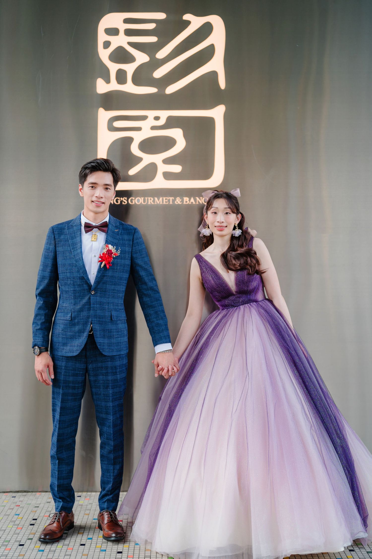 A couple poses in formal attire - checkered blue suit and flowing purple-to-white ombre ballgown - against a neutral background.
