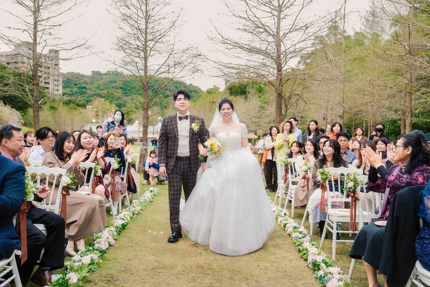 Wedding guests line both sides of an outdoor ceremony aisle decorated with white floral arrangements and chairs.