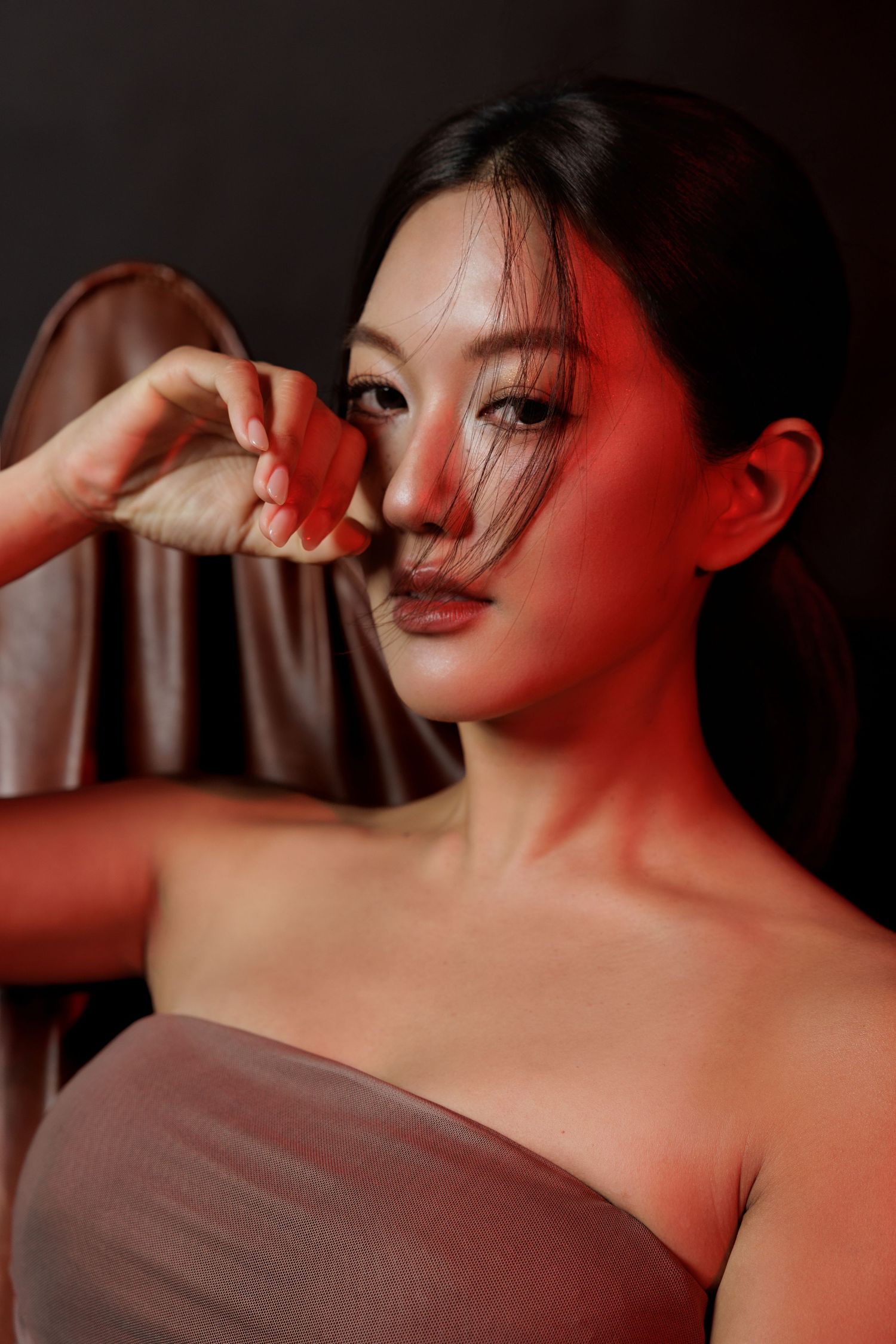 Young Asian woman in strapless dress poses dramatically under red lighting, hand raised near face.