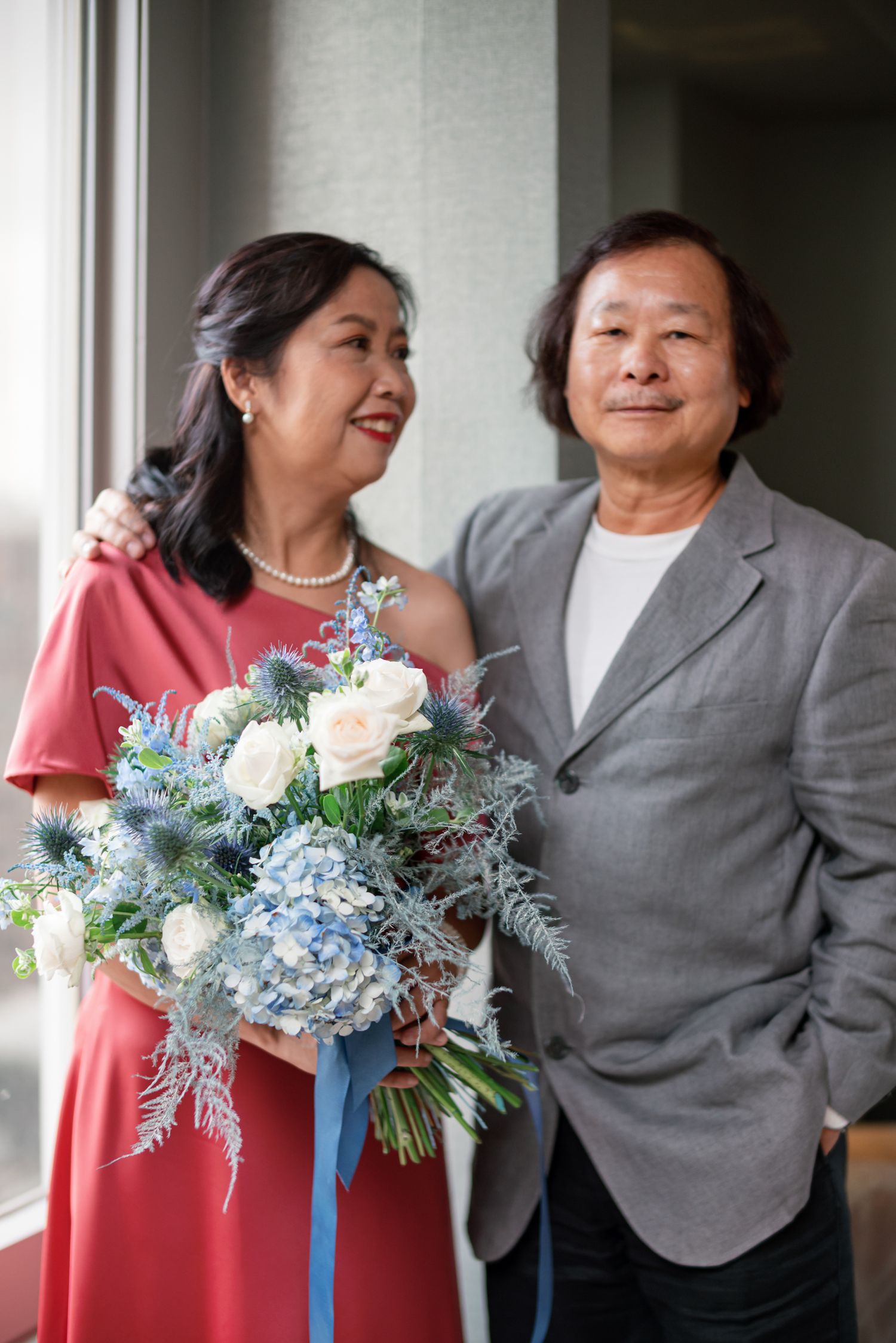 Two people stand together indoors with one holding a beautiful bouquet of blue hydrangeas and white roses with trailing greenery.